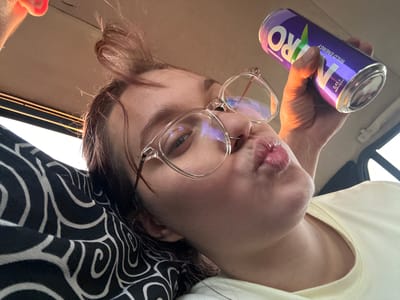 A young woman with light brown hair, wearing clear-rimmed glasses and a light yellow t-shirt, is captured in a close-up selfie. She is holding a purple can of "NITRO WILD ENERGY" in her right hand and puckering her lips. Her left arm rests on a pillow with a black and white swirling pattern. The background suggests an indoor setting, possibly a car, with a beige ceiling and a window visible on the right. The lighting indicates it is daytime, with natural light illuminating the scene. The woman's expression appears playful and relaxed. The visible text on the can reads "NITRO WILD ENERGY," "WILDBERRIES," and "0.45 л". The location context is "Ленин, Kyrgyzstan."