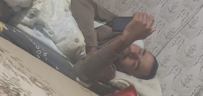 A man is lying on his side, with his eyes closed, holding a dark-colored smartphone in his right hand. He is wearing a light brown shirt. The man appears to be in a relaxed or sleeping state. He is lying on a bed with patterned bedding. The wallpaper in the background also has a floral pattern. A dark gray object with white lettering is visible to the right of the man. The visible text on this object is "60", "b", and some unreadable characters. The lighting suggests it could be daytime, but the specific time of day or weather conditions cannot be determined. The presence of a bed and bedding indicates an indoor setting, likely a bedroom. The provided location context is Konakovo, Russia.