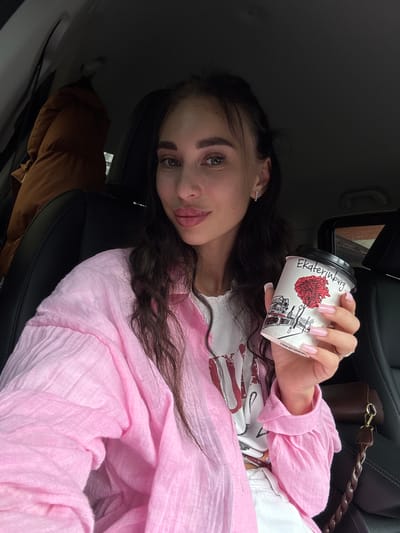 A young woman with dark, wavy hair and striking makeup is seated in the driver's seat of a car. She is wearing a pink, crinkled fabric shirt over a white t-shirt and holding a disposable coffee cup with a picture of a tree and buildings on it. The text on the cup reads "Ekaterinburg". Her fingernails are long and painted a light pink. She is looking at the camera with full lips slightly parted. The car's interior is visible, including a black leather seat and a tan jacket hanging on the back of the passenger seat. The background suggests an outdoor, possibly urban, environment.