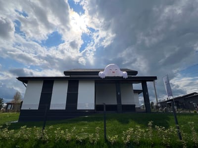 In the city of Yakunino, Russia, a modern, two-story house with a distinctive black and white facade stands under a dramatic sky filled with dark, heavy clouds and patches of bright sunlight. An inflatable cartoon ghost, with large eyes and a smiling mouth, rests on the roof's overhang, appearing to peek over the edge. The property is enclosed by a black metal fence, with a neatly mowed green lawn in the foreground. To the right of the house, a tall flagpole flies a white banner with Cyrillic lettering and a logo, indicating a residential development or real estate agency. Power lines and poles are visible in the background, along with other houses and trees, suggesting a suburban or developing area. The overall scene captures a peculiar blend of architectural design and whimsical decoration against a dynamic weather backdrop.