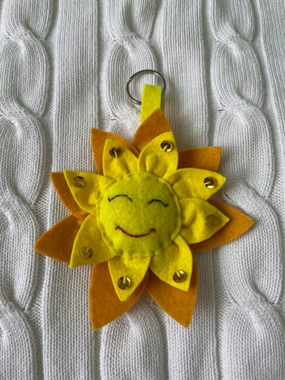 A top-down, eye-level shot shows a yellow and orange felt sunflower key chain with a smiling face on a white knitted blanket. The sunflower has a circular yellow face with black stitched eyes and a small red stitched smile. Gold sequins are attached to the yellow petals. The sunflower has two layers of petals, a yellow inner layer and an orange outer layer. The key chain is attached to the left side of the flower with a small loop of yellow felt. The blanket is a white cable knit pattern. The photograph was taken indoors in Лесной Пейзаж Кп (шарапово, Russia.