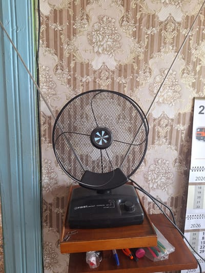 The image shows a close-up of an indoor television antenna with a black circular mesh dish, mounted on a black base. The antenna is positioned on a wooden shelf. To the left of the antenna, a portion of a light blue ribbed surface is visible, possibly a door or wall panel. The background is wallpaper with a floral pattern featuring cream and brown tones. To the right of the antenna, a portion of a calendar for the year 2026 is visible, displaying numbers and some text in Russian. The calendar appears to be a desk calendar with a spiral binding. A few items are on the wooden shelf below the antenna, including paperclips, a plastic bag containing colored pencils or pens, and the tip of a blue pen and orange pencil. The overall scene suggests an indoor setting, possibly a living room or study in a residential area within Блеўчыцкі сельскі Савет, Belarus.