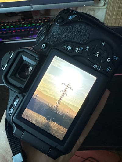 The image shows the back of a black DSLR camera, held by a person whose hand is visible supporting the camera. The camera's LCD screen displays a photograph of a large electricity pylon in a rural landscape at sunset. The sky is filled with warm, golden hues, and the sun is partially obscured by the pylon. The ground appears to be a field with some trees and vegetation. The camera has various buttons and a dial visible, including "MENU," "INFO," "Av," "WB," "SET," and "AF." In the background, out of focus, a computer keyboard with illuminated keys is visible. The setting does not specifically indicate the city of Dachnoye, Russia.