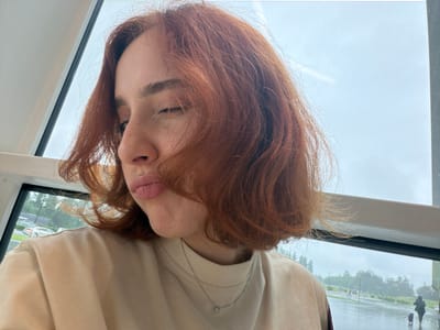 A woman with red hair is visible, her face angled towards the camera. She is wearing a light-colored top and a delicate necklace. She appears to be indoors, looking out through a window. Outside the window, a rainy cityscape is reflected. The location in the city is Ianeti, Georgia. The reflection in the window shows trees and buildings in a soft focus due to the rain. There are people walking with umbrellas on the sidewalk, and vehicles can be seen on the wet roads. The day is overcast.