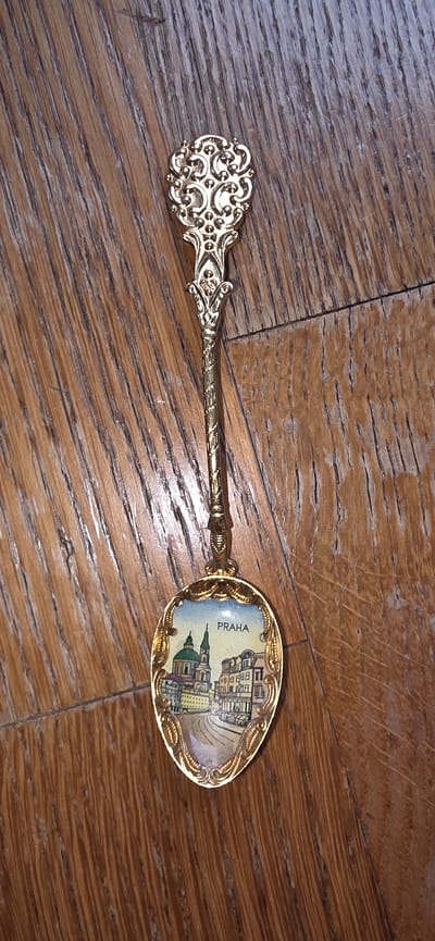 The image is an overhead shot of a decorative spoon lying on a wooden surface. The spoon is gold-colored and features an ornate handle with intricate filigree. The bowl of the spoon is oval-shaped and contains a miniature enamel painting of a European cityscape. The painting depicts buildings with distinct architecture, including a domed church or cathedral with green domes, and other structures with multiple windows and facades. In the background, a bright blue sky with some clouds is visible. The name "PRAHA" is printed in black lettering above the cityscape, indicating that the scene is likely Prague, the capital of the Czech Republic. The wooden surface appears to be made of planks with a visible grain pattern. There is no indication of the city of Marum, Netherlands, in the image.