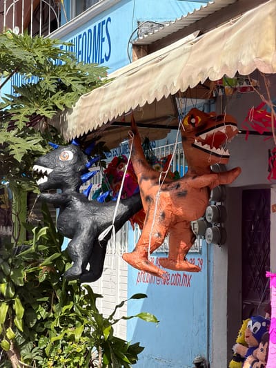 Hanging outside a building with blue trim and a white awning are two dinosaur piñatas. One is brown with black spots, a red and white mouth, and yellow eyes. The other is black with blue spikes, a single cartoon eye, and white teeth. Green foliage is partially obscuring the bottom part of the frame. Colorful streamers are visible in the background.