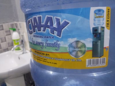 The image shows a close-up of a large blue water dispenser bottle with a label on it. The label has blue and yellow sections. Prominently displayed on the label is the brand name "GWAY" in large blue letters with white stripes. Below this, it says "DRINKING WATER" and "for every family". There is also an image of a smaller water bottle on a water dispenser and text that reads "18.9 Litres". Further down the label, it states "PRODUCED BY: GWAY WATER SCIENCE & TECHNOLOGY CO. LTD." with an address listed as "Ipereway, Isolo, Nigeria". A NAFDAC registration number is also visible. In the background, a white sink and a faucet are partially visible, suggesting the setting is likely a bathroom or kitchen. There are no people or animals in the image. The image is taken from a slightly low angle, focusing on the water bottle's label.