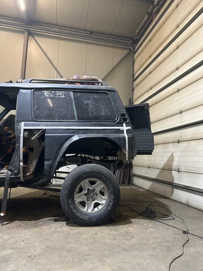 This image depicts the interior of an auto repair workshop located in Musachevo, Bulgaria. The central subject is a dark-colored sport utility vehicle, likely a Nissan Patrol, undergoing body modification or repair. The vehicle is positioned with its passenger side facing the viewer, revealing that the rear quarter panel and fender area have been cut or partially removed. White chalk or masking tape lines are applied to the vehicle's bodywork, indicating planned cut lines for customization.

The vehicle features a roof rack with accumulated dirt or debris, and its rear side windows are dark-tinted or heavily soiled. A white sticker on the rear passenger side window reads "БРАТ ЗА БРАТА" (Brother for Brother). The rear driver's side door is open, and a plaid-patterned fabric is visible on the interior seat. The vehicle is fitted with "COOPER DISCOVERER A/T3" tires on silver-colored alloy wheels.

The workshop environment includes light-colored walls, possibly insulated panels, and a large, beige-colored sectional garage door on the right. Metal structural beams are visible along the walls and ceiling. Overhead fluorescent lighting illuminates the space. The concrete floor is visible, appearing somewhat dusty. An electrical power cord and a small black electrical tool (possibly a grinder or welder) are present on the floor near the rear tire. No individuals are visible in the scene.