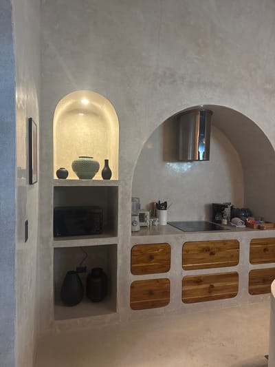This image depicts an interior view of a minimalist kitchen or kitchenette situated in El Nido, Philippines. The space is characterized by light gray, textured concrete or stucco walls and floor. On the left, a built-in vertical structure features two recessed display shelves. The upper shelf, an illuminated arched alcove, showcases three decorative vases: a large, textured teal vase flanked by two smaller, dark-colored vases. Directly below, a black LG microwave oven is situated on a middle shelf, and two dark, bulbous vessels occupy the bottom shelf. A framed artwork hangs on the adjacent wall to the left.
The main kitchen area is set within a large arched concrete alcove, encompassing a gray countertop. A stainless steel cylindrical range hood, bearing the "TEKA" brand, is mounted above a black induction cooktop. To the left of the cooktop, a blender and a white toaster are visible, along with a white utensil holder containing various kitchen tools. To the right, a black coffee maker, several jars, a folded gray towel, and a wooden tray filled with assorted items are arranged on the counter. Below the countertop, the concrete base incorporates six wooden cabinet doors, each with rounded corners and a central circular pull. No individuals are present in the scene.