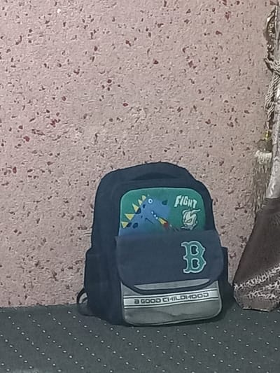 A navy blue backpack with a dinosaur theme rests diagonally on a textured surface. The backpack's front features a green patch with a cartoon dinosaur and the word "FIGHT." A flap with a stylized "B" covers part of the green patch. The side of the backpack has a gray panel with the phrase "A GOOD CHILDHOOD." The surface beneath the backpack is a speckled beige, transitioning to a polka-dotted gray at the right edge. A portion of a patterned object appears above the backpack.