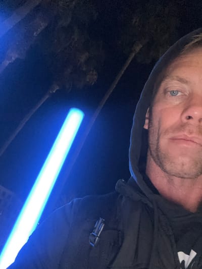 The image captures a nighttime selfie of a male individual in Long Beach, United States of America. The subject is depicted from mid-face to upper chest, possessing light blue eyes and visible stubble. He wears a dark hooded sweatshirt, with the hood pulled over his head. A metallic-colored buckle or clip is visible on the right shoulder of his garment or a strap worn over it. To the left and slightly behind the subject, a prominent, elongated cylindrical object emits an intense, bright blue light, consistent with the appearance of a prop lightsaber. The background is dark, indicating an outdoor nighttime setting, with the silhouettes of multiple tall palm trees faintly discernible. A indistinct lighter structure is visible in the lower left background, beneath the glowing blue object.