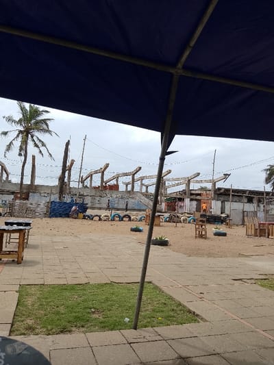 The image shows a view from under a blue tarp canopy, looking out towards a paved area with a sandy ground and a large, incomplete concrete structure in the background. Several wooden tables with chairs are scattered throughout the paved area, with some tire-like objects scattered in front of them. There are four figures in the distance, including one wearing an orange shirt. The structure behind is a skeletal framework of an incomplete building, perhaps a stadium, with numerous concrete beams running across a large open space. There are power lines running across the scene. The scene is in Lomé, Togo, near the coordinates latitude: 6.169, longitude: 1.353. The environment is dry and dusty with a clear sky. There is a large palm tree visible in the distance to the left.
