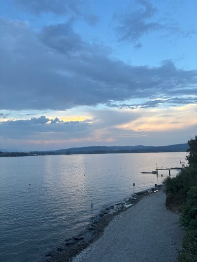 The image depicts a serene lake scene at dusk, likely taken from a slightly elevated viewpoint overlooking a shoreline. The sky is a mix of deep blues and lighter hues of pink and yellow, with scattered clouds, suggesting the sun has recently set or is about to rise. The lake's surface is calm, reflecting the colors of the sky, with gentle ripples indicating a light breeze.

In the foreground, a pebble beach transitions into a rocky embankment. A signpost stands on the rocky edge. Further out in the water, several yellow buoys are visible. To the right, a wooden pier extends into the lake, with a small structure at its end. Dense green foliage and trees line the upper right portion of the frame, with a lifeguard chair visible near the pier. The opposite shore is visible in the distance, with a line of hills or mountains forming a hazy backdrop. The overall impression is one of tranquility and natural beauty.

The provided context suggests the location is the city of Arona, Italy, which is situated on the shores of Lake Maggiore.