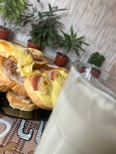 The image displays a close-up, slightly tilted view of several open-faced sandwiches or toasts arranged on a dark, round plate. The food is the central focus, featuring slices of light-colored bread topped with melted, golden-yellow cheese. Some slices also include round pieces of reddish-brown sausage or cured meat beneath the cheese, while others have a spread of what appears to be minced meat or a pâté-like mixture under the melted cheese.
In the right foreground, a clear glass or pitcher, partially obscured, holds an opaque, milky-white liquid, likely milk. The background is softly blurred, revealing an indoor setting. To the left, several green potted plants are visible. Behind the plants, a light-colored wall features a textured pattern resembling bricks or tiles in white and grey tones. A patterned placemat or tablecloth in brown and beige tones, adorned with abstract and geometric designs, including houndstooth-like motifs, is visible beneath the plate and to the right.
The scene depicts a prepared meal or snack, likely consumed in a home environment such as a kitchen or dining room. No people are visible, and there is no discernible text within the image. The time of day cannot be determined. The location context for this media file is Ботево, Bulgaria.