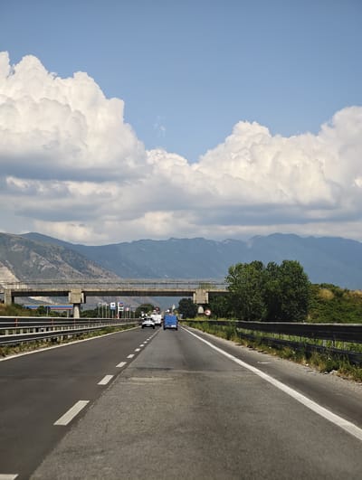 The image depicts a highway scene with mountainous terrain in the background. A multi-lane highway stretches towards the horizon, bordered by guardrails. Several vehicles are visible on the road, including cars and a van. An overpass crosses the highway in the middle ground. The sky is partly cloudy with large, fluffy white clouds against a blue backdrop. The overall impression is a sunny day in a rural or semi-rural setting. This highway is located in or near the city of Acerra, Italy.