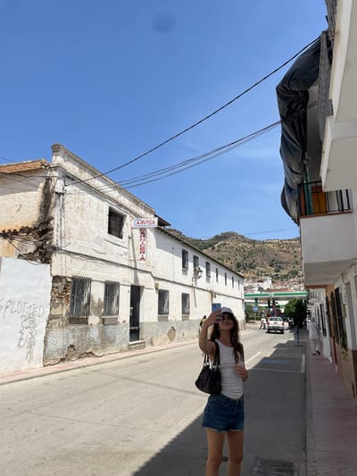 A young woman with long brown hair wears a white tank top, denim shorts, sunglasses, and a white baseball cap. She stands on a paved street in Pizarra, Spain, holding a blue smartphone and taking a selfie. She carries a black shoulder bag. The street is lined with buildings, some appearing older and weathered with exposed brick and peeling paint, others more modern with white facades. A sign reading "AMPISA CARNICERIA" is visible on one building. In the background, a BP gas station with green and white branding is situated on a hill. More buildings and hills with sparse vegetation stretch into the distance under a clear blue sky. Power lines crisscross the sky above the street.