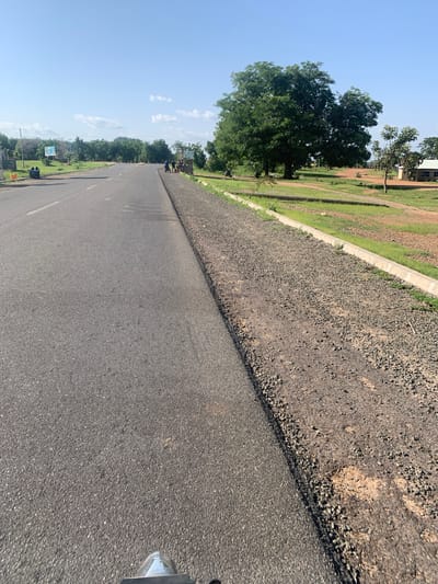 The media file depicts a wide shot of a freshly paved asphalt road, angled to show the perspective of someone standing on or near it. The road surface is dark gray and appears smooth, with a white line marking the lane. To the left of the road, there's a raised embankment covered in dark gravel, separating the asphalt from a grassy area. Further left, lush green trees and a clear blue sky with scattered white clouds dominate the scene. The time of day appears to be late morning or early afternoon, with bright sunlight casting shadows.

In the background, glimpses of buildings and more trees suggest a semi-urban or rural setting, possibly a town or community in Boi A, Nigeria. A few people are visible in the distance along the roadside, some appearing to be walking or standing near a sign. The overall impression is one of ongoing infrastructure development or maintenance, with the new road being a focal point. The weather seems pleasant and sunny. There is no visible text in the image.