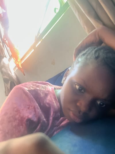 A close-up, upward-angled shot depicts a person with dark skin and dark hair. The person is wearing a pink shirt. Sunlight is visible through a window, creating a bright glare in the lower left of the frame. The location is inferred to be Onibudo, Nigeria. Details on any actions, interactions, or other subjects are not observable from the available data.