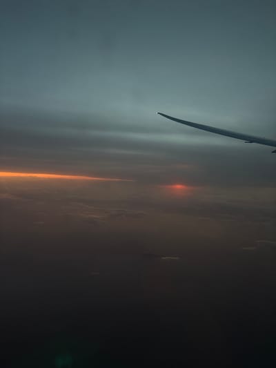 The image depicts an aerial view from an aircraft, likely at high altitude during either sunrise or sunset. The right wing of the aircraft is visible in the upper-right quadrant. The sky transitions from a muted blue-gray at the top to dark grey layered clouds. A prominent horizontal band of orange light is visible mid-left, above a dense cloud layer, while a more intense, diffused red glow is present mid-right, possibly indicating the sun's position. A vast, multi-layered cloudscape obscures the entire lower half of the frame, preventing any ground or water features from being observed. The overall illumination is low. A faint green lens flare is present in the lower-left corner. No people or specific actions are depicted. The image lacks any identifiable geographical features necessary to determine its location within the Cotswold District, United Kingdom; the view is entirely above extensive cloud cover. Cotswold District is a rural area, not a city.