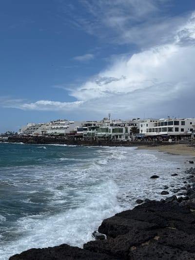 The image depicts a daytime coastal scene in Playa Blanca, Spain, observed from a low vantage point on the shoreline. The foreground consists of dark, irregular volcanic rock formations along the water's edge on the right, transitioning into turbulent turquoise-blue ocean water with white-capped waves breaking towards the shore.
Mid-ground, a strip of light-colored sand forms a small beach to the left of the volcanic rocks, with several small human figures visible on it and in the shallow water. Beyond the sandy area, the coastline becomes rocky again, supporting a continuous line of multi-story, white-plastered buildings that define the waterfront. These structures feature dark-framed windows and balconies, with many ground-floor units displaying green awnings or storefronts, indicative of commercial establishments such as restaurants and shops. A paved promenade runs along the base of these buildings, populated by numerous individuals walking or congregating, and outdoor seating areas are visible. A single palm tree stands out among the buildings towards the center-right.
The background shows a partially cloudy sky. The upper left quadrant features clear blue sky, while the upper right and central sections are filled with large, diffuse white and grey cumulus clouds. A small, dark, airborne object, possibly a bird, is discernible in the upper left clear sky. Rooftops of the buildings display various utilities, including a visible antenna mast.
