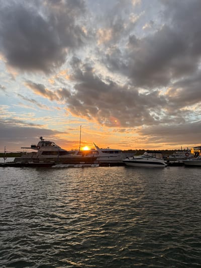 In the city of Molodezhnyi, Russia, a serene sunset casts a warm glow over a harbor filled with moored yachts. The sky is a canvas of dramatic clouds, tinged with hues of orange and pink, as the sun dips below the horizon. The water reflects the sky's colors, creating a shimmering effect. Several yachts of varying sizes are docked, their elegant lines silhouetted against the vibrant sky. The scene captures a tranquil moment in the harbor, a peaceful end to the day.
