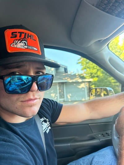 The scene, set in Rancho Murieta, United States, depicts a young adult male in the driver's seat of a vehicle, looking directly at the camera. He wears a black baseball cap featuring an orange rectangular patch with the white "STIHL" logo and a chainsaw illustration. Black square-framed sunglasses with blue mirrored lenses cover his eyes. He is dressed in a dark blue short-sleeved t-shirt with a partially visible light-colored graphic on the left chest, including text and the year "2024," and blue jeans. A gray seatbelt crosses his chest. His right hand is positioned on the dark-colored steering wheel. The vehicle's interior includes a dark dashboard, door panel, and a light beige headliner. Through the vehicle's windows, a residential street is visible. In the background, there is a two-story house with a stone facade on the lower level and a lighter-colored upper level, along with a garage door. Green deciduous trees are prominent behind and to the right of the house, against a clear blue sky. The reflection in the sunglasses shows additional interior elements of the vehicle. The overall lighting suggests daytime conditions.