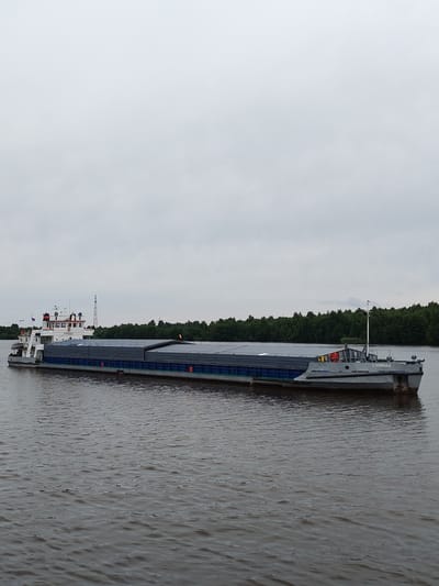 A barge is seen in the water, with a two-story cabin structure at the front, painted white with red and black accents, and bearing the Cyrillic inscription "СИММАХ". The barge is loaded with a long, rectangular cargo cover, dark gray in color, with blue trim along the bottom edge. In the background, a dense line of green trees forms the shoreline, and a tall, lattice-like structure, possibly a power line pylon, is visible against a cloudy sky. The water is choppy, with ripples on its surface. The scene appears to be in a rural or semi-rural area along a waterway.