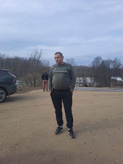 The scene depicts an outdoor setting in Avdot’ino, Russia, under an overcast sky. In the foreground, a Caucasian male stands in the center-right on an unpaved, light brown surface. He has short dark hair and a neutral expression. He is wearing a dark olive crew-neck sweatshirt with white text on the chest, black athletic-style pants, and dark athletic shoes with white soles. His hands are in his pockets, and a watch is visible on his left wrist.

To the left of the frame, the rear driver's side of a dark-colored SUV is visible, parked on the same unpaved surface. Further into the background, slightly to the left and behind the vehicle, a second person, appearing female, is partially visible from behind. This individual is wearing a dark hooded jacket and light-colored patterned pants, facing away from the camera.

The background features numerous bare deciduous trees, indicating late winter or early spring conditions. Beyond the trees, a body of water, partially covered with ice or snow, is discernible.