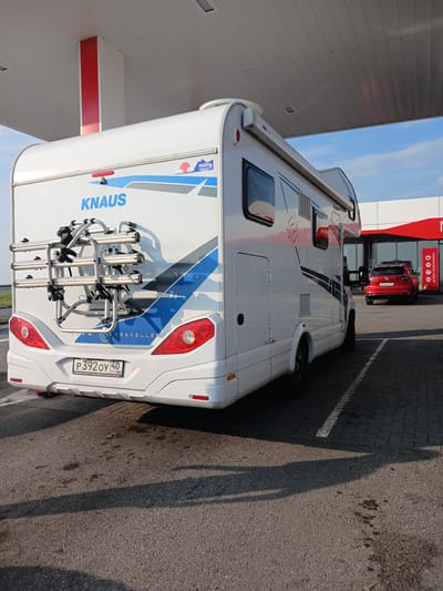 A white KNAUS caravan is parked under a covered structure, likely at a gas station or service area, on a paved surface. The caravan has a bike rack mounted on the rear, holding two bicycles. The license plate is visible and reads "P 392 OY 40 RUS". In the background, a red Volkswagen SUV is parked near a building with red accents and glass doors. The environment appears to be a roadside service area in Nazitamak, Russia, indicated by the language on the building signage and the Russian license plate. The sky is blue with some clouds, and the sun is casting shadows.
