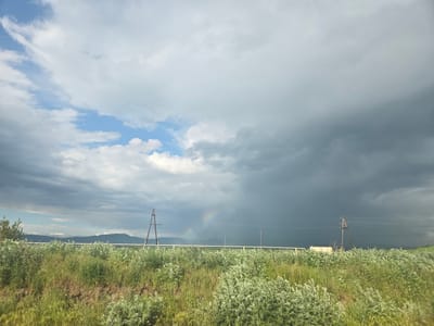 This is a landscape photograph taken in the city of Azatan, Armenia. The image features a wide, green field in the foreground, dotted with various types of vegetation and some patches of dry earth. In the mid-ground, there's a white horizontal fence, and behind it, several wooden utility poles are visible, supporting power lines that stretch across the frame. Beyond the fence and poles, dark, ominous storm clouds dominate the sky, contrasting with patches of lighter, brighter clouds and a sliver of blue sky on the left. A faint rainbow is visible in the distance, arching across the stormy sky. In the far background, a range of dark mountains forms the horizon. The scene appears to capture the dramatic atmosphere just before or during a rain shower. There are no people or animals visible in the image.