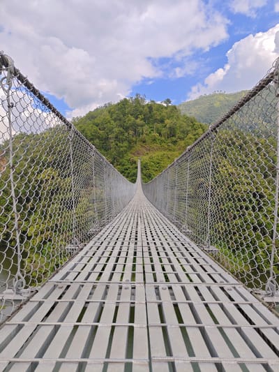 A wide-angle, eye-level shot captures the perspective from the middle of a long, metal suspension bridge in Lekhnath, Nepal. The bridge deck is made of parallel metal slats with gaps between them, revealing a body of water below. Chain-link fencing runs along both sides of the bridge, extending towards the horizon. Beyond the fencing, lush green hills covered in dense trees rise up, meeting a sky with scattered white clouds against a blue backdrop. The perspective of the bridge draws the viewer's eye towards a distant point where the bridge seems to disappear into the landscape. The image is taken during daylight, with natural lighting illuminating the scene. There are no people or animals visible in the frame. The overall scene depicts a natural landscape with human-made infrastructure.