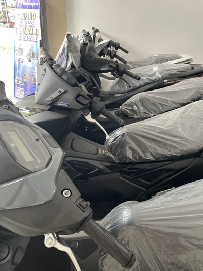 A row of motorcycles, covered in protective plastic, are displayed inside a shop. In the foreground, the front section of a gray motorcycle is visible, featuring its handlebars and a digital display. To the right, a black motorcycle is partially seen, also covered in plastic. Further back, more motorcycles are lined up. A blue and yellow banner, likely an advertisement, is visible on the left side of the image, with text in Filipino. The banner mentions "Transcycle" and what appears to be pricing information, with one price at "₱3,800". The motorcycles are arranged in a showroom setting, with a light-colored wall in the background. The scene appears to be in San Jose del Monte, Philippines, as indicated by the text on the banner.