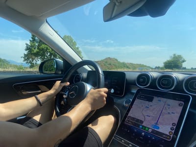 The image shows the interior of a car, viewed from the passenger seat, looking towards the driver and the road ahead. A person is driving, with their hands on the steering wheel. The car's dashboard is visible, featuring a large touchscreen display showing a navigation map. The map indicates a route with "Uscita Brescia Est" (Exit Brescia East) labeled, suggesting the location is near Brescia, Italy. The road outside appears to be a highway or a similar type of road, with green trees and a slightly hilly landscape visible under a clear blue sky. The overall impression is a snapshot of a journey, with the focus on the driving experience and the navigation system. The specific location, Azzano San Paolo, Italy, is not explicitly visible or indicated within the image itself, but the presence of "Uscita Brescia Est" on the navigation screen strongly suggests a location within the vicinity of Brescia, which Azzano San Paolo is a part of.
