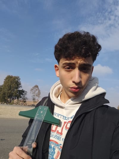 A young male, appearing to be in his late teens or early twenties, is depicted in an outdoor setting in Mankat al-Hatab, Syria. He possesses curly dark brown hair and light brown eyes, looking directly at the camera with a neutral facial expression and slightly parted lips. He is dressed in a light-colored hooded sweatshirt, partially revealing a "WESTERN POLO" graphic print, beneath an unzipped black puffer jacket. A black backpack strap is visible over his left shoulder. The subject holds a T-square in his right hand; the tool features a green, T-shaped head and a transparent plastic blade with a black strap or attachment. The background features a clear blue sky with scattered white cirrus clouds. In the mid-ground, an asphalt road or path is visible. Beyond the road, the landscape consists of arid, rocky ground with sparse vegetation, including a large, leafy green tree on the left and thinner trees in the distance. The scene is brightly lit by natural daylight.