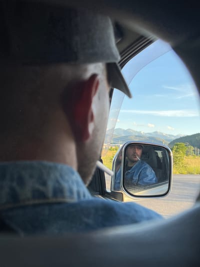 A man wearing a baseball cap and a denim jacket is reflected in a side-view mirror of a vehicle. He is smoking a cigarette. The background of the mirror shows a landscape of mountains and trees under a clear blue sky. The man's back and part of his head are visible in the foreground, suggesting the photo is taken from inside the vehicle, looking out. The image is taken in Dragushinovo, Bulgaria.
