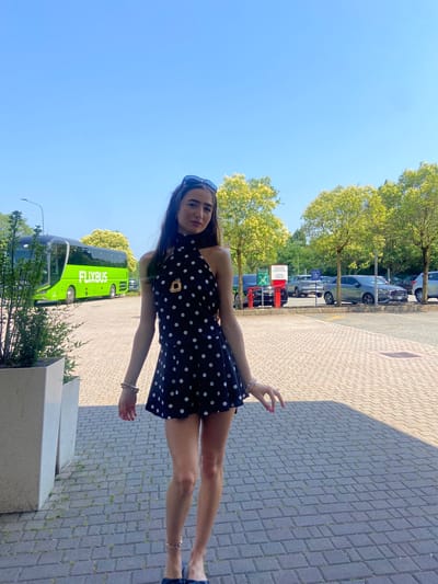 A young woman stands in a paved outdoor area, looking towards the camera. She is wearing a black sleeveless dress with white polka dots, a necklace with a gold pendant, and sunglasses perched on her head. The woman has long, dark hair and is adorned with bracelets on her left wrist and an anklet on her right ankle. She is positioned in front of a green FlixBus coach. In the background, several cars are parked, and trees with light green foliage line the area. The ground is paved with rectangular interlocking bricks. The scene suggests a transportation hub or a rest stop, likely near the Malpensa Airport in Somma Lombardo, Italy, given the FlixBus presence and typical infrastructure for such a location. The sky is clear and blue, indicating a sunny day.
