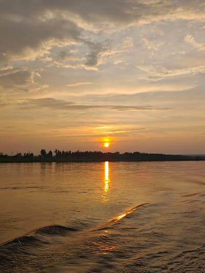The image depicts a serene sunset over a body of water, likely a river or lake, near Maloe Nakhval'skoe, Russia. The sun is low on the horizon, casting a warm, golden glow across the scene. The sky is filled with soft, wispy clouds tinged with orange and yellow hues. A dark, silhouetted treeline forms a distant horizon. The water is calm, with gentle ripples reflecting the sunset's light. The foreground shows small waves and the wake of a boat, suggesting movement through the water. No people or buildings are visible in the frame. The overall atmosphere is tranquil and picturesque.