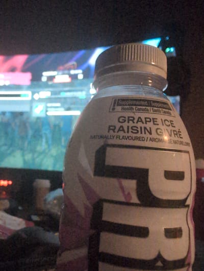 The image shows a close-up of a bottle of "PRIME Hydration" drink, flavored "Grape Ice Raisin Givré." The bottle is primarily white with pink and purple graphic elements. Text on the bottle indicates it is "Naturally Flavoured / Aromatisé Naturellement." The bottle's cap is ribbed and light-colored. In the background, out of focus, is a television screen displaying colorful graphics, possibly from a video game or a streaming service. To the left and slightly behind the bottle, a blurred image of a coffee cup is visible. The overall setting appears to be an indoor environment, likely a living room or recreational space, given the presence of the television and casual atmosphere. There are no people directly visible in the frame, but the scene suggests a person was likely holding the bottle or in the vicinity of the setup. The location in Kingston, Canada, is not discernible from the visual information provided in the image.