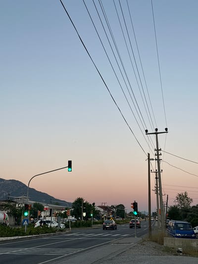 The image depicts a scene in Köyceğiz, Turkey, at dusk. The sky transitions from light blue to a pale orange. A series of power lines and utility poles run across the top and left of the frame. A road runs along the right of the image with cars and various parked vehicles. Several traffic lights are visible, all displaying green signals. Bushes and other foliage line the roadside. A mountain range is visible in the lower portion of the image.