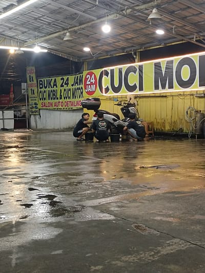 This is a vertical, medium-shot, eye-level photo taken at a motorcycle and car wash in Kelapa Dua, Indonesia. The scene is lit by overhead fluorescent lights, giving it a slightly eerie, nighttime feel. The ground is wet and reflective, suggesting recent rain or ongoing washing.

In the foreground, the wet concrete floor dominates, with puddles and streaks reflecting the lights above. In the middle ground, three young men are washing a motorcycle. Two of them are kneeling by the rear of the bike, actively cleaning it, while a third stands slightly behind. They are wearing dark t-shirts, one with a "TOP" logo. A motorcycle is parked, partially obscured by the men, and a few buckets are scattered on the wet ground.

A large, bright yellow banner with black text stretches across the top of the scene. The text, "BUKA 24 JAM CUCI MOBIL & CUCI MOTOR SALON AUTO DETAILING CUCI MOL", advertises the business as being open 24 hours for car and motorcycle washing and detailing. Another smaller sign to the left lists "CUCI 8X GRATIS 1X SEMARIN HEMAT", indicating a loyalty program.

To the far left, a portion of a red and white striped cooler or vending machine with "PUCUK" branding is visible, along with a black tarp. The overall atmosphere is utilitarian, with the focus on the service being provided. The time of day appears to be evening or night, given the artificial lighting and the activity.