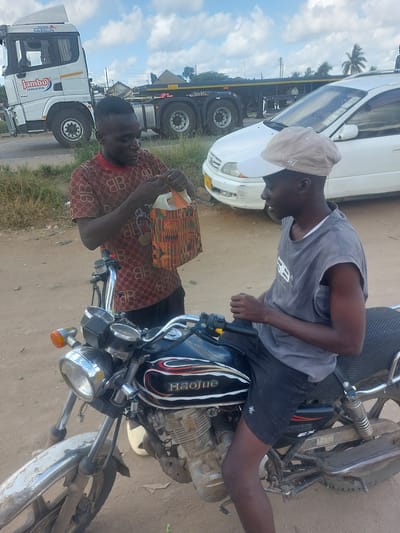 In Picha Ya Ndege, Tanzania, two young men are interacting near a motorcycle and a white car. One man, wearing a maroon t-shirt with a repeating pattern, stands by the motorcycle, holding a colorful woven bag with a plastic handle and an orange top. The other man, wearing a grey sleeveless shirt and a light-colored cap, is seated on the motorcycle, facing the first man. The motorcycle appears to be black and silver, with the "Haojue" brand visible on its side. In the background, a large white truck with "JAMBO JAMUKAYA" printed on it is parked, along with another trailer. The environment is dusty and appears to be along a roadside, with dry grass and vegetation present. The sky is partly cloudy.
