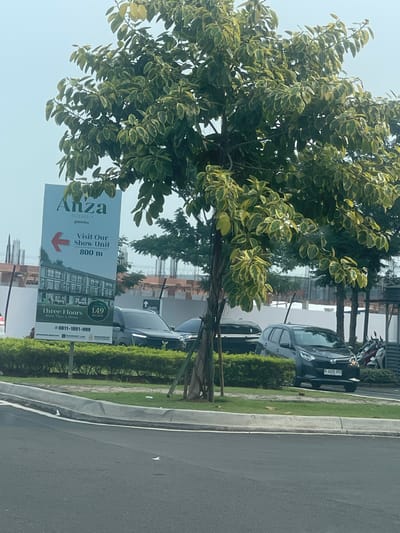 The image depicts a street scene in Cijantra, Indonesia, with a prominent billboard advertising "Anza Homes @Malibu." The billboard, a tall, rectangular sign with a blue and white design, features an illustration of a three-story building, the text "Three Floors More Than a Home," and information about a price of "149 BILLION." It also includes a phone number: "0811-1891-999," and logos for "PARAMOUNT LAND." An arrow points left, indicating to "Visit Our Show Unit 800 m."

In front of the billboard, a lush green hedge runs along the side of the road. Behind the hedge, several vehicles are parked or driving. A dark-colored SUV is visible on the right, with a license plate reading "B 1220 RYJ." To its left, a dark-colored sedan is parked, and further left, another vehicle is partially obscured. Several motorcycles are also parked behind the SUV.

A large tree with variegated green and yellow leaves dominates the foreground, its branches extending across the top of the image. The tree has a thick, dark trunk that appears to be supported by a wooden stake. In the background, construction is underway, with visible scaffolding and unfinished buildings. The sky is a pale blue, suggesting an overcast or hazy day. The overall scene appears to be an entrance to a residential development.