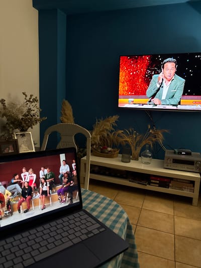The image captures an indoor setting, likely a living room, within Urbanización Encinares De Alcolea, Spain. The scene features a large, wall-mounted flat-screen television on a dark teal wall, displaying a man in a light green suit jacket, appearing animated against a background of sparkling orange and red lights. Below the TV, a white shelving unit holds an audio system, books, and various decorative items including dried plants and vases. To the right, a laptop rests on a surface covered with a blue and white checkered blanket or tablecloth, its screen showing a group of people seated in what appears to be a classroom or audition setup.
The primary activity depicted is simultaneous media consumption: someone is watching a program on the large television and engaging with content on the laptop. The man on the TV seems to be a presenter or performer in a live broadcast.
Notable details include the contrasting dark teal wall with a lighter upper section and ceiling, light brown tiled flooring, and ambient lighting that suggests evening or nighttime. The decorative elements, such as dried grasses, multiple vases, and framed pictures near the floor, contribute to a homey atmosphere.
Visible text includes "DIRECTO" (Spanish for "LIVE") on the large TV screen, a small "HD" icon, and potentially a channel logo like "LA 1". On the audio system, "gms." is visible. The spine of a book on the shelf clearly shows "FOLLETT".