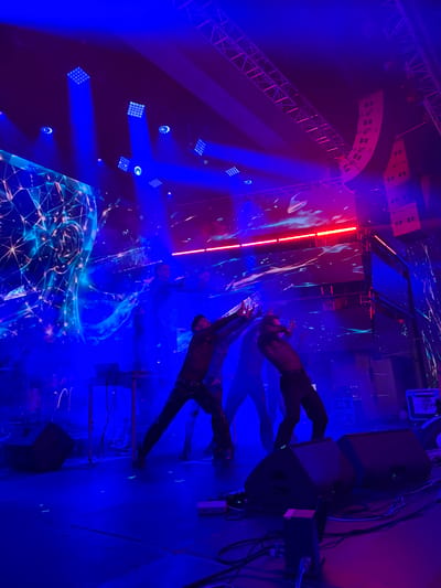 The image captures a dimly lit stage with performers, likely a dance or music event, bathed in vibrant blue and pink theatrical lighting. The stage features large LED screens displaying abstract, glowing visuals. Several individuals are visible in various dynamic poses, suggesting an active performance. Stage equipment, including speakers and a table with a laptop, are present. The overall atmosphere is energetic and modern, with a focus on visual spectacle. The image was taken in Tsilkani, Georgia.