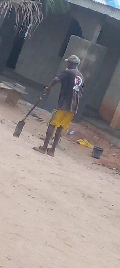 A person is shown wielding a shovel, likely cleaning or moving soil in Agunmo, Nigeria. The location has dry, dusty ground. A bucket and yellow cloth are nearby. The person wears a dark t-shirt, yellow shorts, and a hat. An unfinished building is behind them.