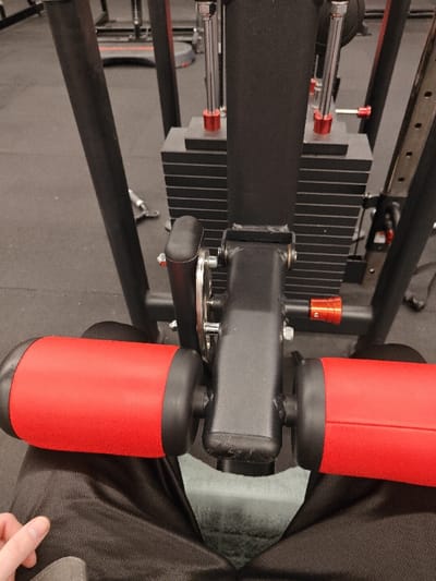 The image shows a person seated at a weight machine. The machine has red padded bars for leg support. The weight stack is black, with red accents. A black arm and adjustable orange knob are also visible. The person wears dark pants. The background consists of a gray floor and indistinct gym equipment.