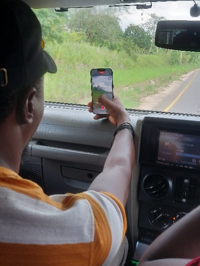 The scene is captured from within a vehicle, looking forward, reportedly in Kisanga, Tanzania. In the foreground, the upper back and right side of a person are visible, seated in what appears to be the passenger seat. The individual wears a black baseball cap with a yellow element and an off-white and orange horizontally striped shirt. A black bracelet adorns their right wrist. Their right hand holds a dark-colored smartphone upright, its screen displaying an active camera interface mirroring the exterior view. A red recording icon and a red circular button, presumably for camera operation, are visible on the phone's screen, with the person's thumb positioned near it. The vehicle's interior shows a grey dashboard and a black console unit with air vents and a digital display showing "BLUETOOTH Audio" and "21:40". Beyond the windshield, a paved road with a yellow shoulder line extends into the distance, flanked on both sides by dense green grass and lush trees, characteristic of a rural or undeveloped natural environment. The sky is largely obscured by foliage but appears muted.