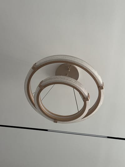 A modern, circular chandelier hangs from a textured white ceiling. The chandelier consists of two interlocking rings, each with a golden frame and a translucent, ribbed interior that emits a soft light. The rings are suspended by thin black wires. Below the chandelier, a dark, horizontal line suggests a partition or a shelf. The overall environment appears to be an indoor space, likely a room in Ынтымак, Kyrgyzstan, characterized by its clean, minimalist design and subdued lighting. There are no people or visible actions in the frame.