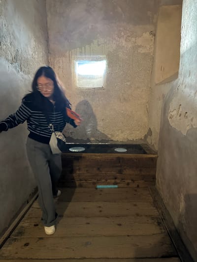 The image shows a person standing in a stone room with a small window. The room appears to be old, with textured walls and a wooden door on the right. The person is wearing a black and white striped shirt, gray pants, and white shoes. The picture was taken in the city of Veytaux, Switzerland.