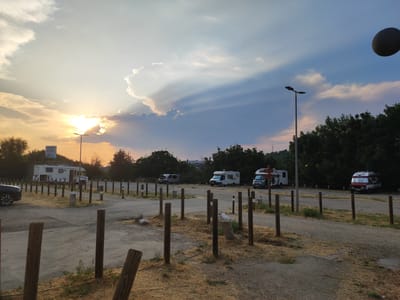 The image depicts a campsite at dusk, likely located in Béjar, Spain. The sun is setting, casting a warm glow and dramatic light rays through the clouds. Several recreational vehicles (RVs) are parked in an open, dusty area marked by wooden posts. In the background, a line of trees forms a dense border. A sign that reads "CAMINO NATURAL" is visible on a pole, indicating a natural path or trail, which is characteristic of rural or semi-rural Spanish landscapes. The overall atmosphere is serene, with the fading sunlight creating a peaceful and tranquil scene. There are no people or active interactions visible. The ground is covered in gravel and dry grass, typical of a campsite setting. The sky is a mix of blues, grays, and oranges due to the sunset.